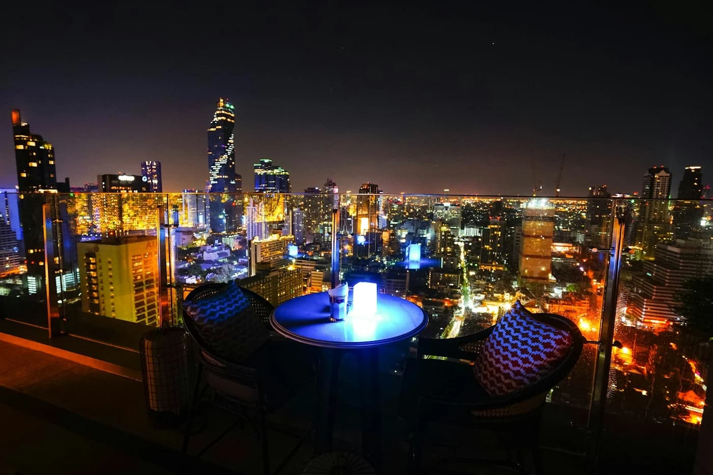 A rooftop table and chairs beside a glass railing, overlooking Bangkok’s illuminated skyline.