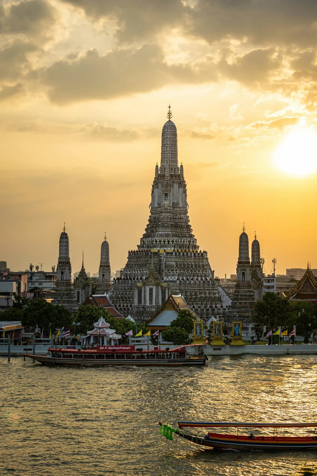 Wat Arun rising above the Chao Phraya River beneath golden clouds, with longtail boats in the foreground.
