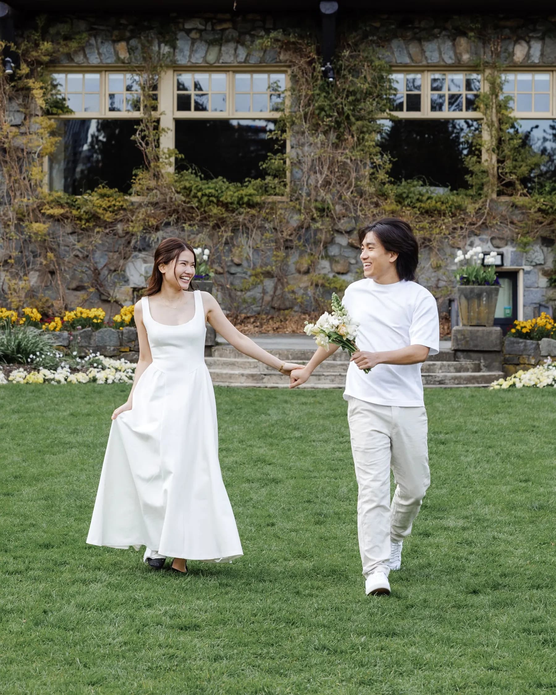 Jam and Tony walking hand in hand across a lawn, both in white, laughing at each other, an ivy-covered stone lodge behind them