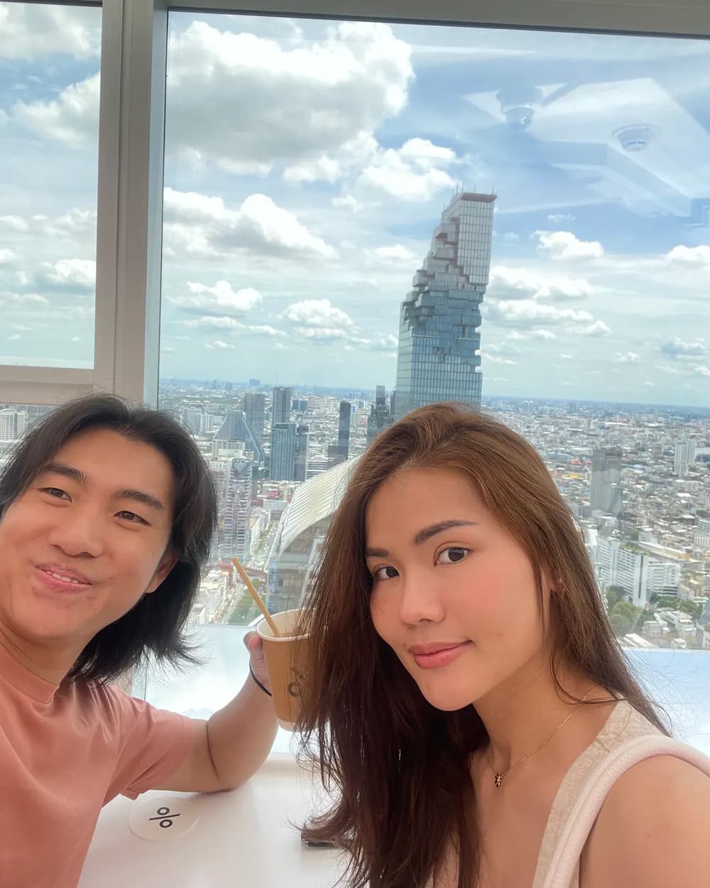 Jam and Tony with iced coffee at a high window, the pixelated MahaNakhon tower filling the skyline behind