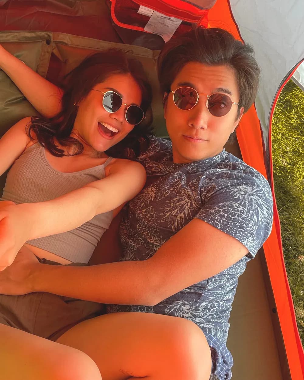 Jam and Tony lying side by side inside an orange tent, Jam laughing, both in sunglasses