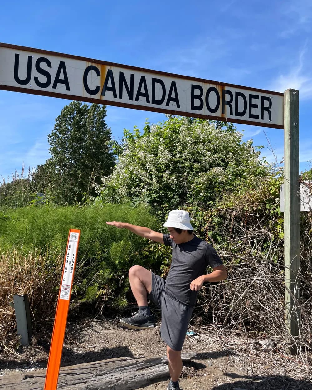 Tony striking a dramatic pose under a wooden sign reading USA CANADA BORDER, an orange boundary marker beside him