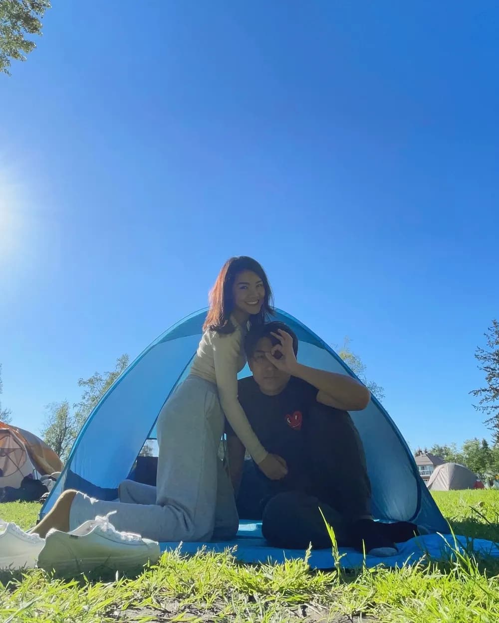 Jam and Tony under a blue pop-up tent on a sunlit lawn, other tents pitched across the grass around them