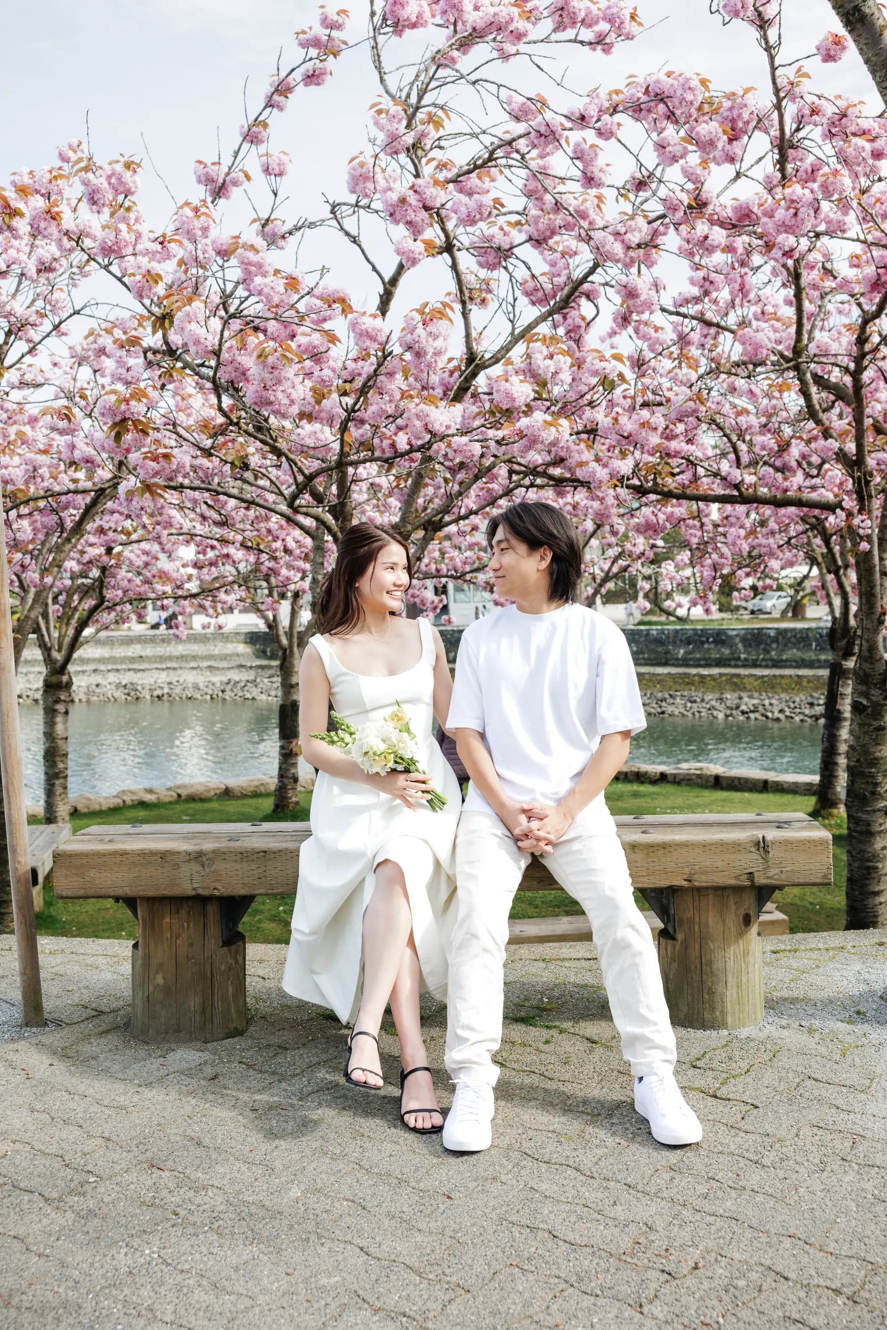 Sitting together on a wooden bench under cherry blossoms by the water