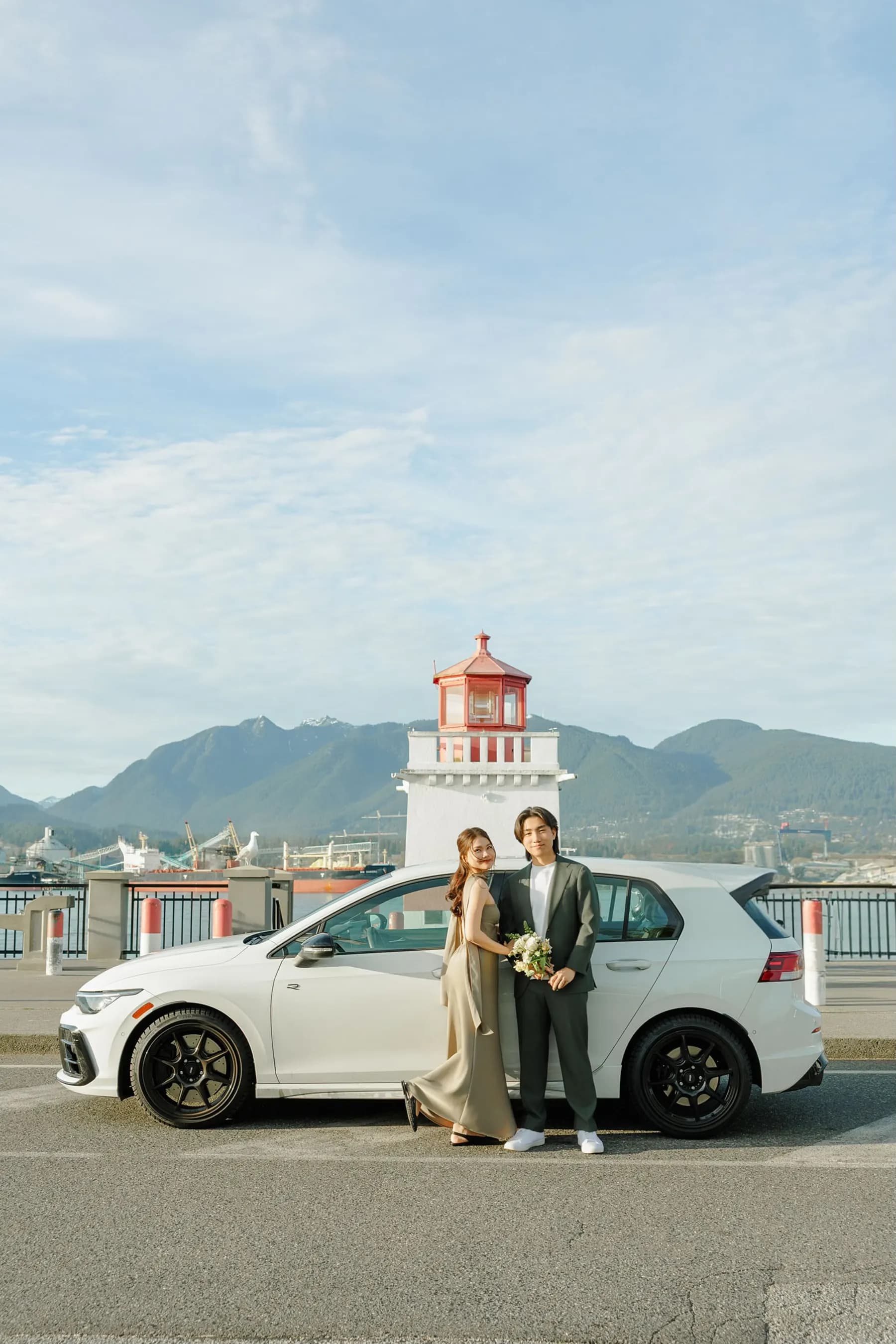 Standing together beside their white car, bouquet in hand, the red-topped Brockton Point lighthouse and the North Shore mountains behind